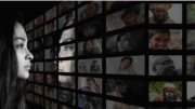 Photo of a person in black and white with long hair looking at different people through a variety screens in front of them.
