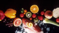 Image of red fish, red, green, and orange fruits and vegetables, and a slice of bread on a black background.