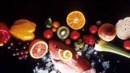 Image of red fish, red, green, and orange fruits and vegetables, and a slice of bread on a black background.