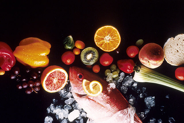 Image of red fish, red, green, and orange fruits and vegetables, and a slice of bread on a black background.