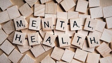 A series of wooden Bananagram letters spelling "MENTAL HEALTH" on a background of blank tiles.