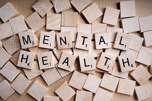 A series of wooden Bananagram letters spelling "MENTAL HEALTH" on a background of blank tiles.